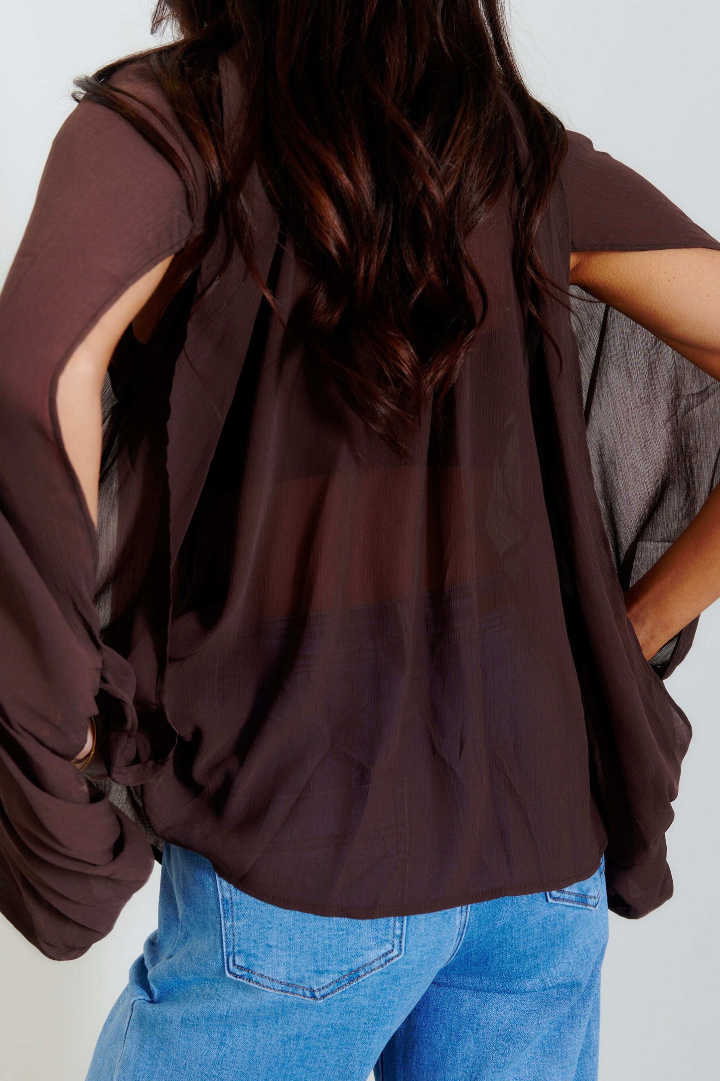 A woman wearing a dark brown sheer long-sleeve top with side cutouts and blue jeans, shown from the back against a light gray background.