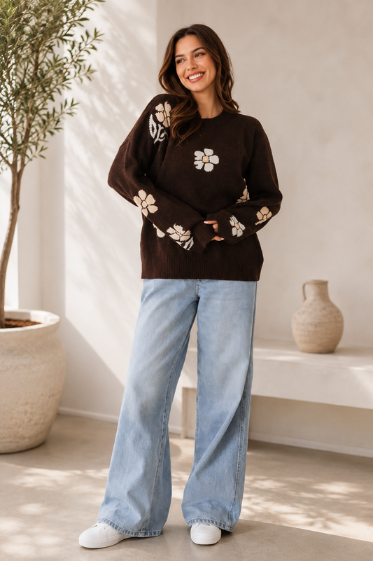 A woman wearing a dark brown crewneck sweater with white floral embroidery, paired with light-wash wide-leg jeans and white sneakers, standing in a brightly lit room with a potted plant and a small vase in the background.