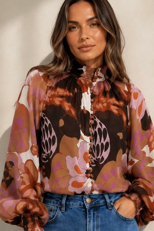 A woman wearing a sheer long-sleeve blouse with a bold floral and abstract print in brown, orange, pink, and black, paired with blue jeans against a neutral beige background.