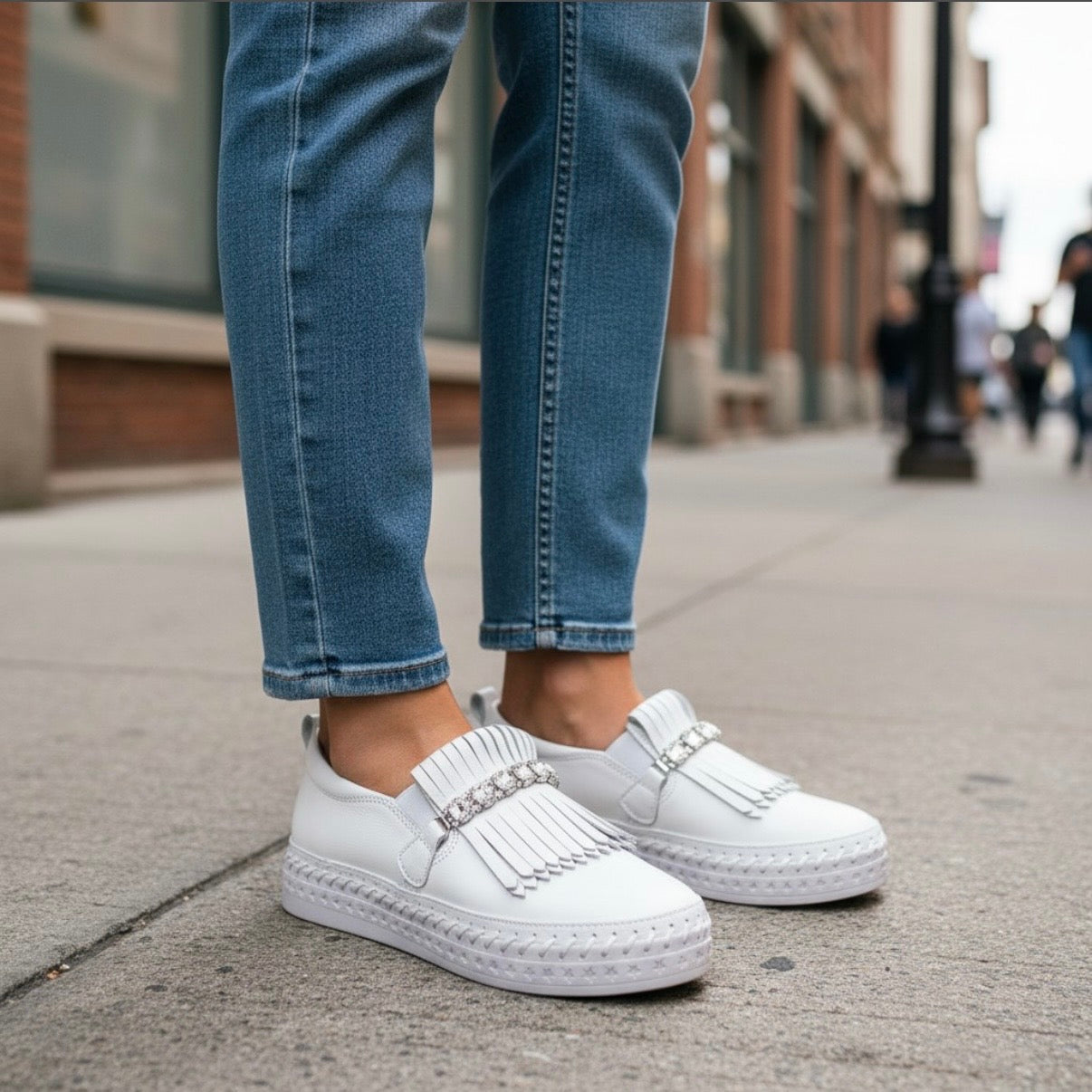 White slip-on platform sneakers with fringe and rhinestone trim, shown on a person wearing blue jeans on a city sidewalk.