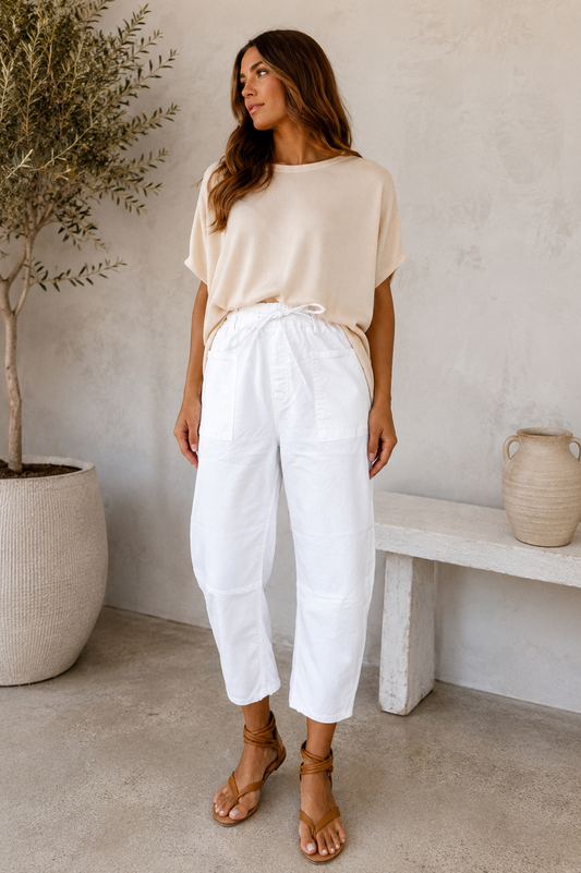A woman modeling a beige short-sleeve t-shirt and white drawstring cropped pants, paired with tan strappy sandals, standing in a minimalist, neutral-toned room with a potted olive tree and a stone bench.