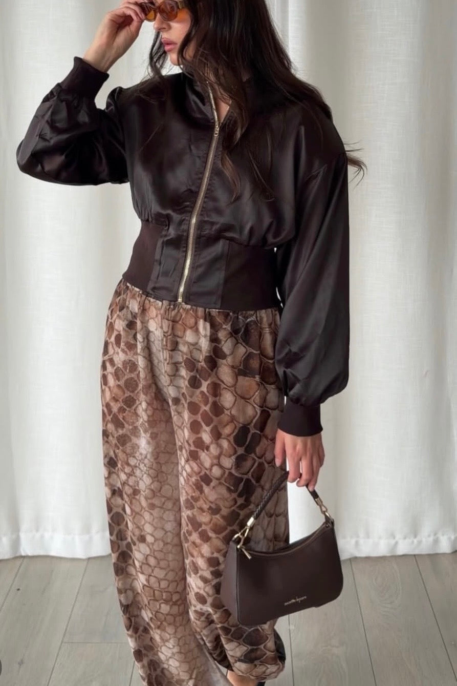 A woman wearing a dark brown satin bomber jacket and brown snakeskin print pants, holding a dark brown handbag against a white curtain background.