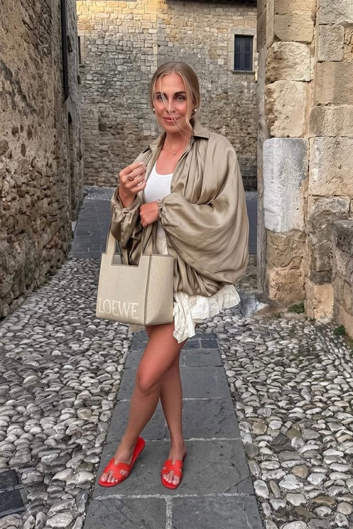 A woman stands in a narrow cobblestone alleyway wearing a beige satin-like draped jacket over a white top and white skirt, carrying a beige tote bag with 'LOEWE' printed on it, and wearing red flat sandals.
