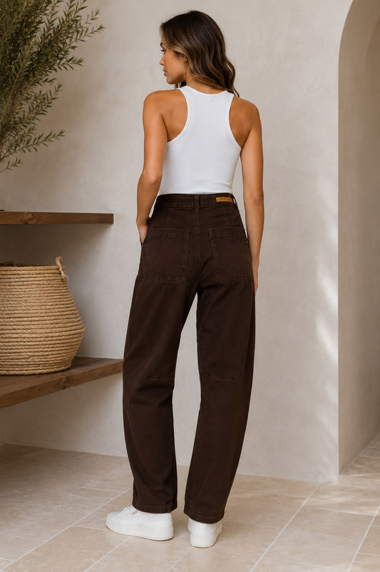 A woman shown from the back wearing dark brown wide-leg pants, a white ribbed tank top, and white sneakers in a warm, minimalist interior setting.
