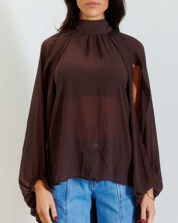 A woman wearing a sheer dark brown mock-neck blouse with long balloon sleeves and a gathered neckline, paired with blue jeans, against a plain light gray background.