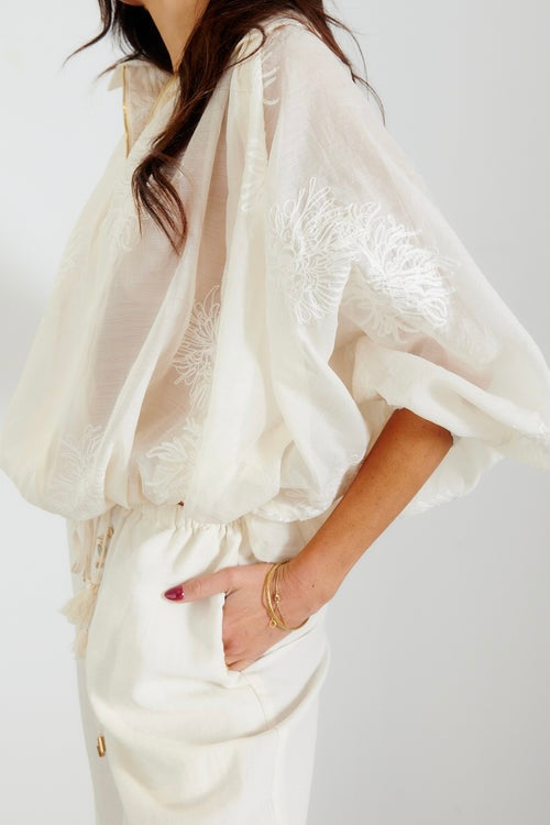 White semi-sheer embroidered batwing top with collared V-neckline and drawstring waist, styled with matching cream wide-leg pants.