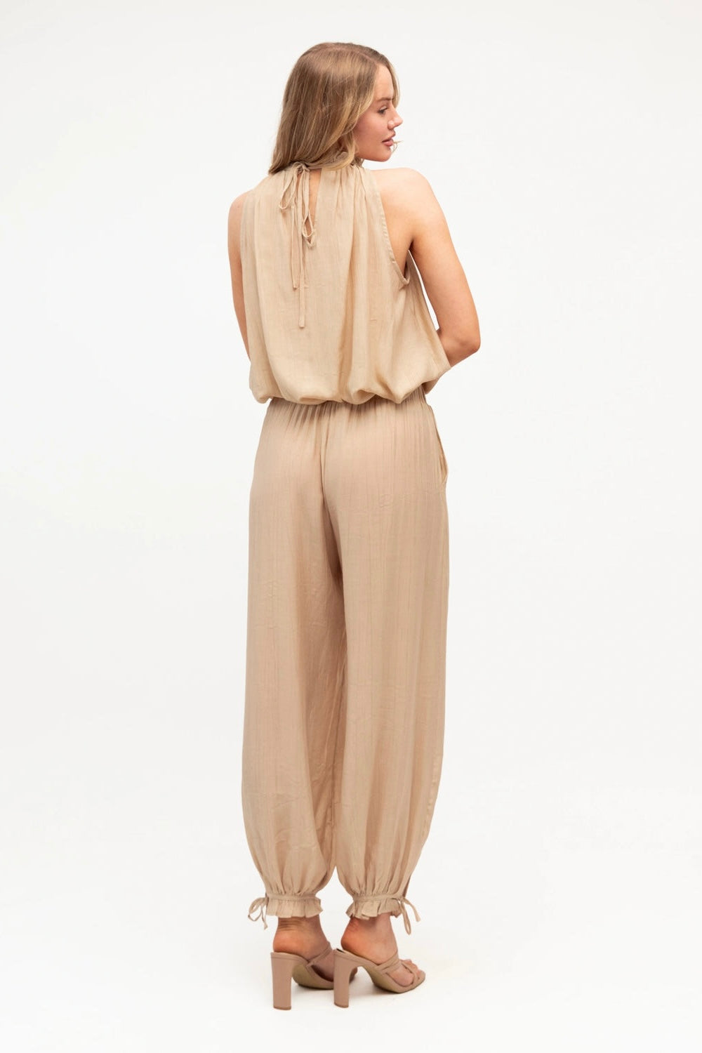 A woman shown from the back wearing a light beige sleeveless halter jumpsuit with gathered fabric and elasticized ankle ties, paired with beige heeled sandals, against a plain white studio background.