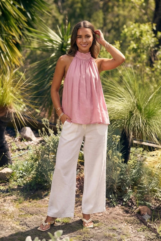 A woman stands outdoors in a garden setting wearing a sleeveless pink halter top and white wide-leg pants.