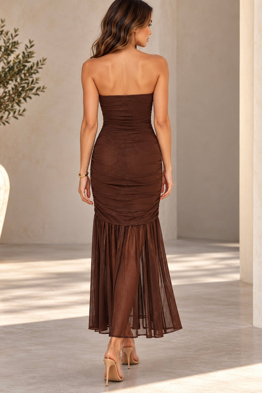 A woman wearing a strapless ruched maxi dress in dark brown, shown from the back as she walks in a sunlit architectural setting with beige walls and columns.