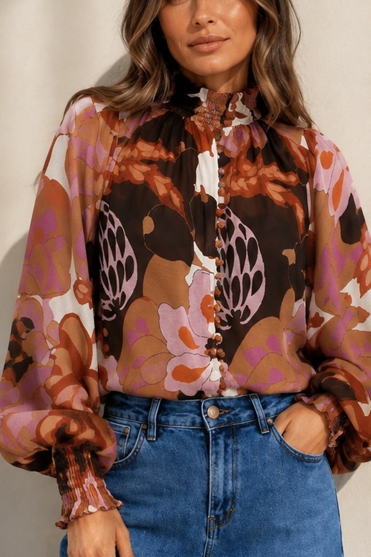 A woman wearing a long-sleeve high-neck blouse with a bold retro floral and abstract print in brown, pink, orange, and white, paired with blue jeans.