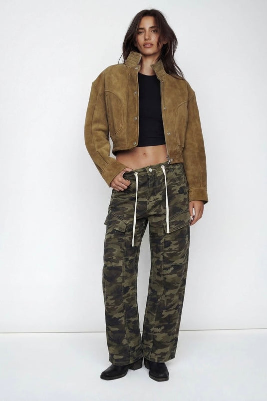 A model stands against a plain white studio background wearing a cropped brown suede jacket, a black cropped top, and wide-leg camouflage cargo pants with white drawstrings.