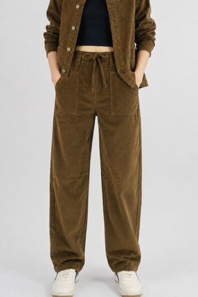 A person wearing wide-leg brown corduroy pants with a drawstring waist, styled with a dark top, brown jacket, and white sneakers against a light gray studio background.