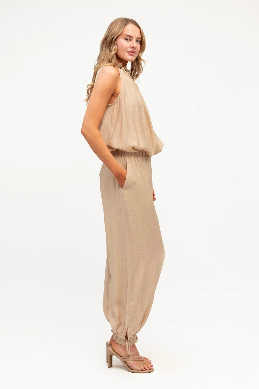 A woman modeling a sleeveless beige jumpsuit with a draped top and wide, baggy harem-style legs, paired with matching heeled sandals, against a plain white studio background.