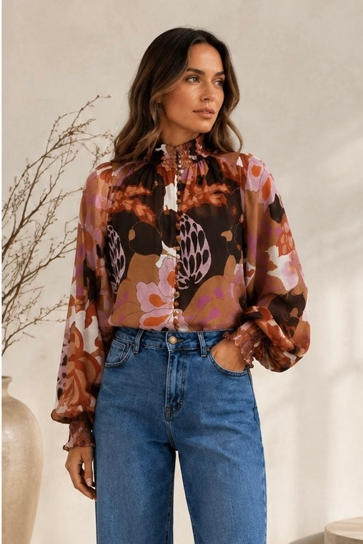 A woman wearing a long-sleeve high-neck blouse with a brown, pink, and beige floral pattern, paired with blue jeans, standing against a neutral beige wall.