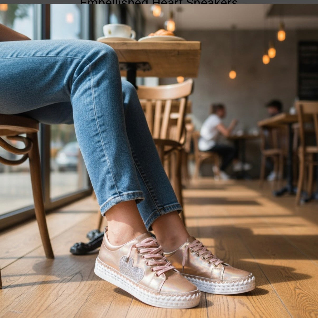 A person seated in a cafe wearing metallic rose gold sneakers with pink laces and blue jeans, shown from the thighs down in a warm, sunlit indoor setting.