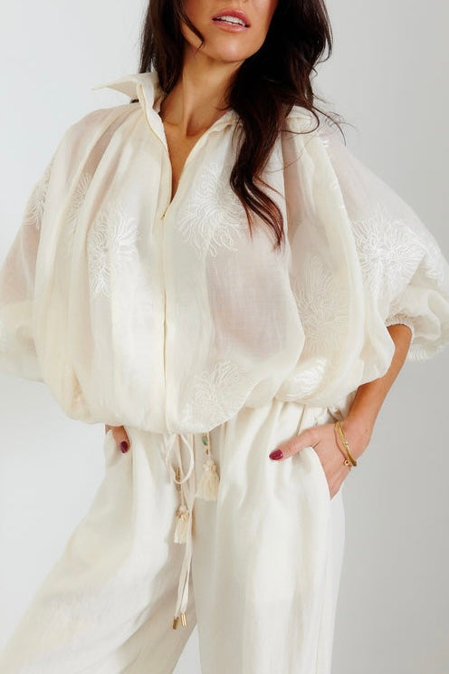 A woman wearing a cream-colored sheer embroidered blouse with voluminous balloon sleeves and matching cream wide-leg trousers, shown against a light gray studio background.