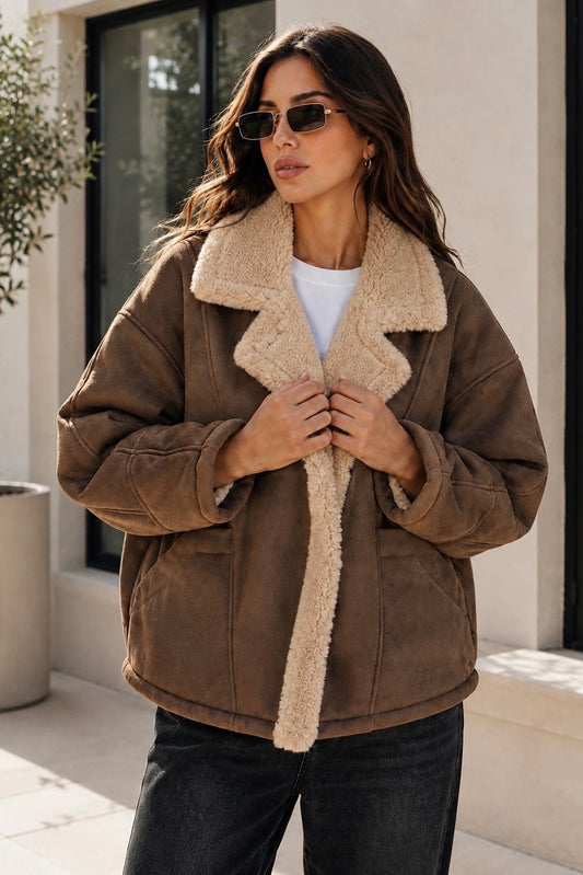 A woman wearing a brown suede-like jacket with a thick beige sherpa collar and lining, styled with a white t-shirt and dark jeans.