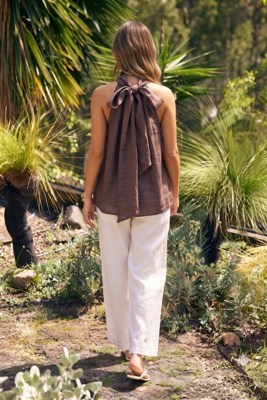 A woman viewed from behind walking outdoors on a garden path, wearing a sleeveless brown top with a large bow detail at the back and white wide-leg pants.
