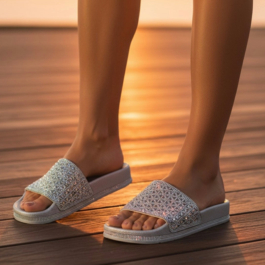 A close-up of a person wearing sparkling silver slide sandals with rhinestone-embellished straps, standing on a wooden deck at sunset.
