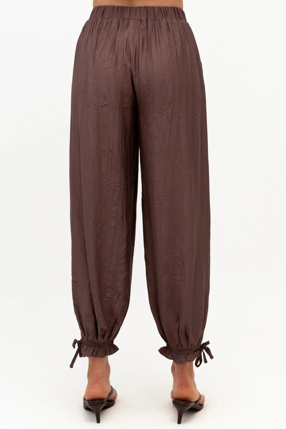 A pair of loose, dark brown crinkled trousers with an elastic waistband and gathered ankle ties, shown on a model from the back against a white background.