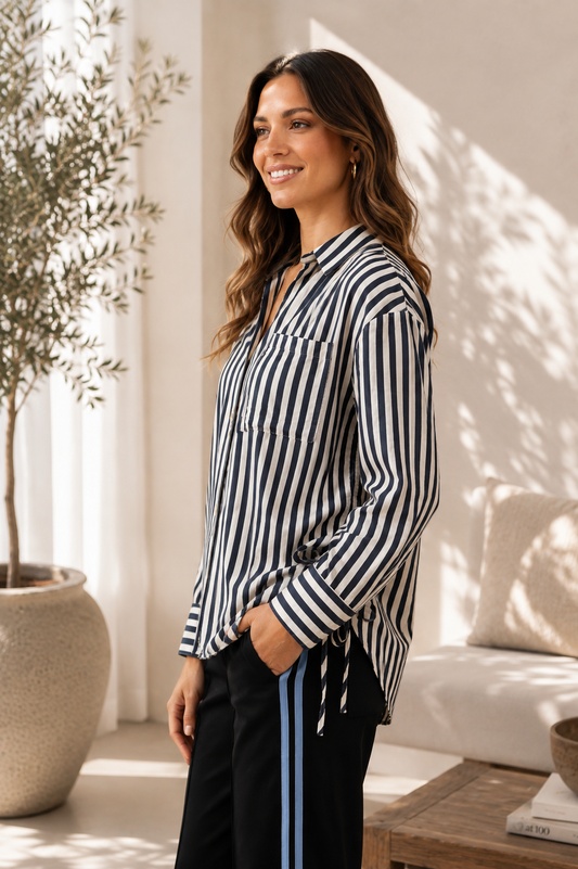 A woman wearing a black and white vertically striped long-sleeve blouse with a split neckline and drawstring detail, paired with black pants featuring a blue side stripe, standing in a brightly lit, modern living room.