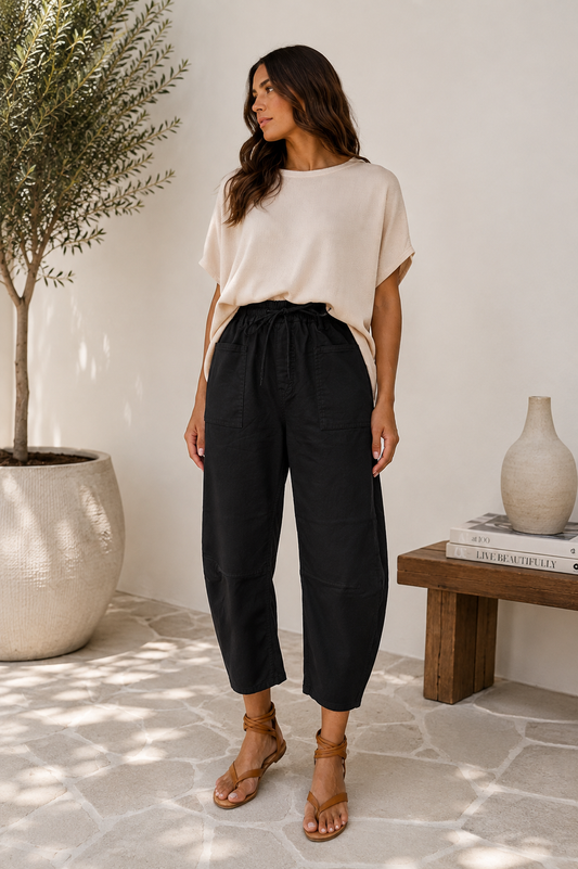 A woman stands in a bright indoor setting wearing a cream short-sleeve top and black high-waisted cropped pants with brown strappy sandals. A potted tree, a small wooden table with a vase, and a stone floor are visible in the background.