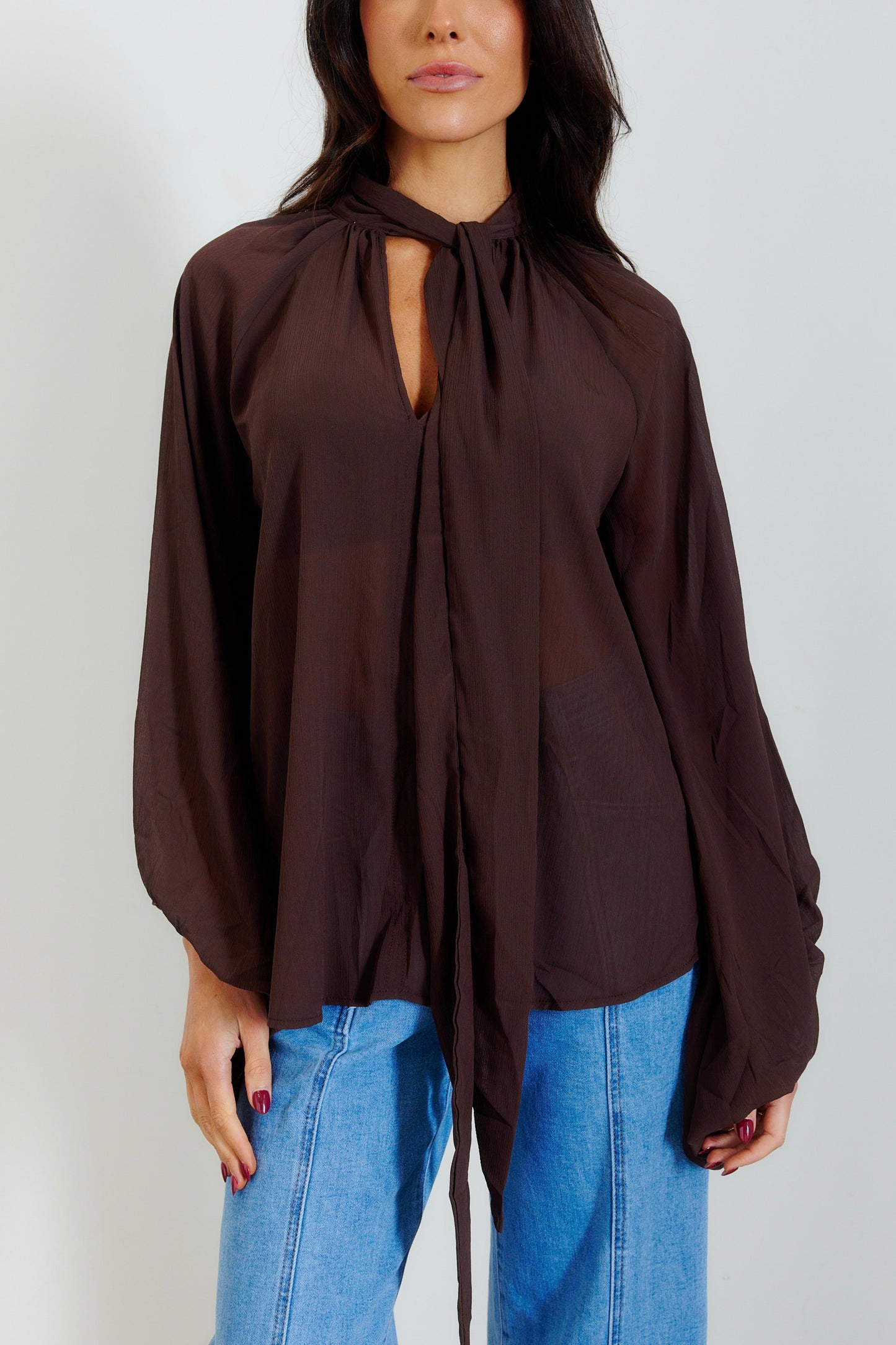 A woman wearing a dark brown long-sleeve blouse with a keyhole tie-neck detail and blue jeans, shown from the nose down against a plain light gray background.