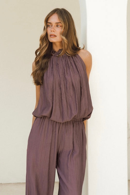 A woman modeling a mauve-purple sleeveless jumpsuit with a gathered high neckline and wide-leg pants, standing against a clean white architectural background.