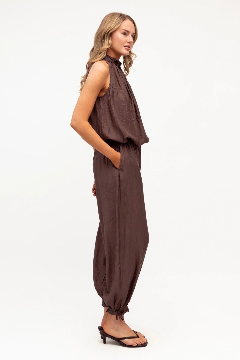 A woman modeling a sleeveless dark brown jumpsuit with a gathered, draped top and loose, balloon-style legs, shown in profile against a plain white studio background.