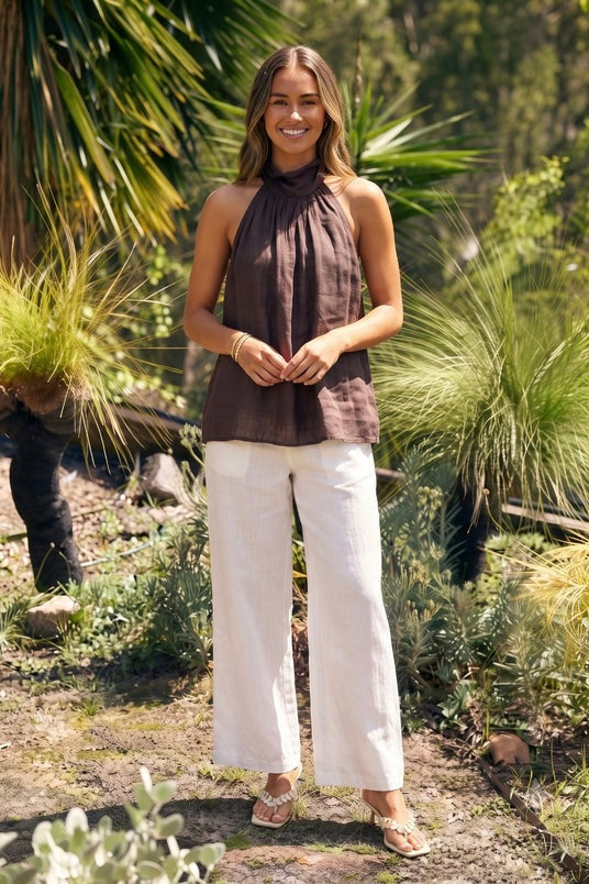 A woman stands outdoors in a garden setting wearing a sleeveless brown halter top and white wide-leg cropped pants.