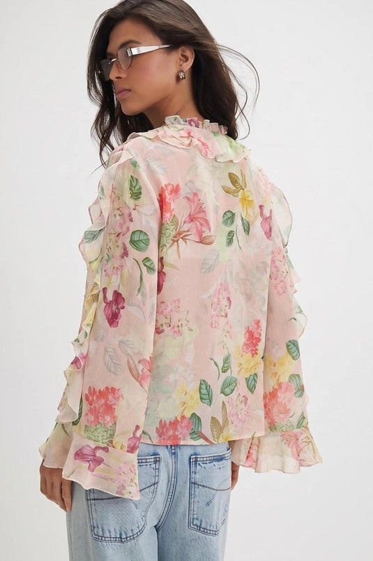 A woman wearing a light pink long-sleeve blouse with a colorful floral print and ruffled trim, paired with light blue jeans, shown from a side-back angle against a plain white background.