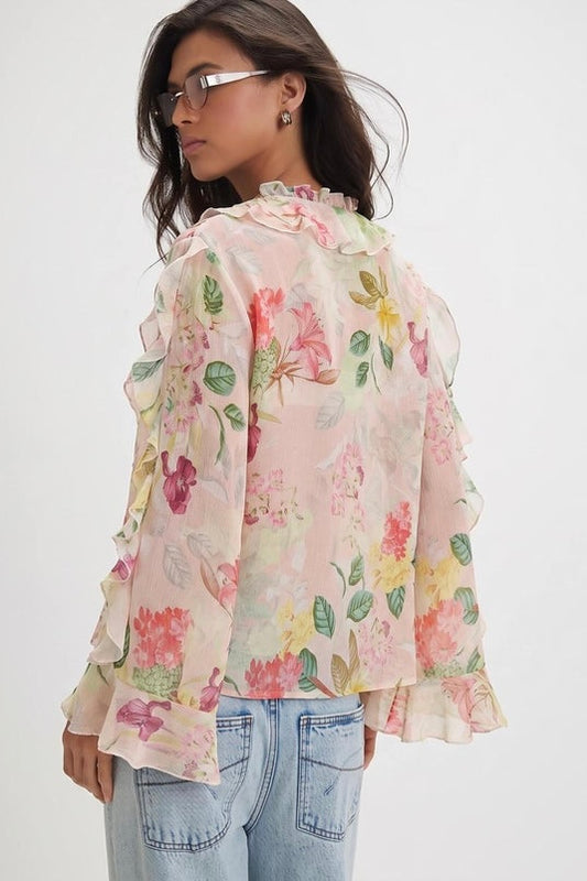 A woman wearing a light pink long-sleeve blouse with a colorful floral print and ruffled trim, paired with light blue jeans, shown from a side-back angle against a plain white background.