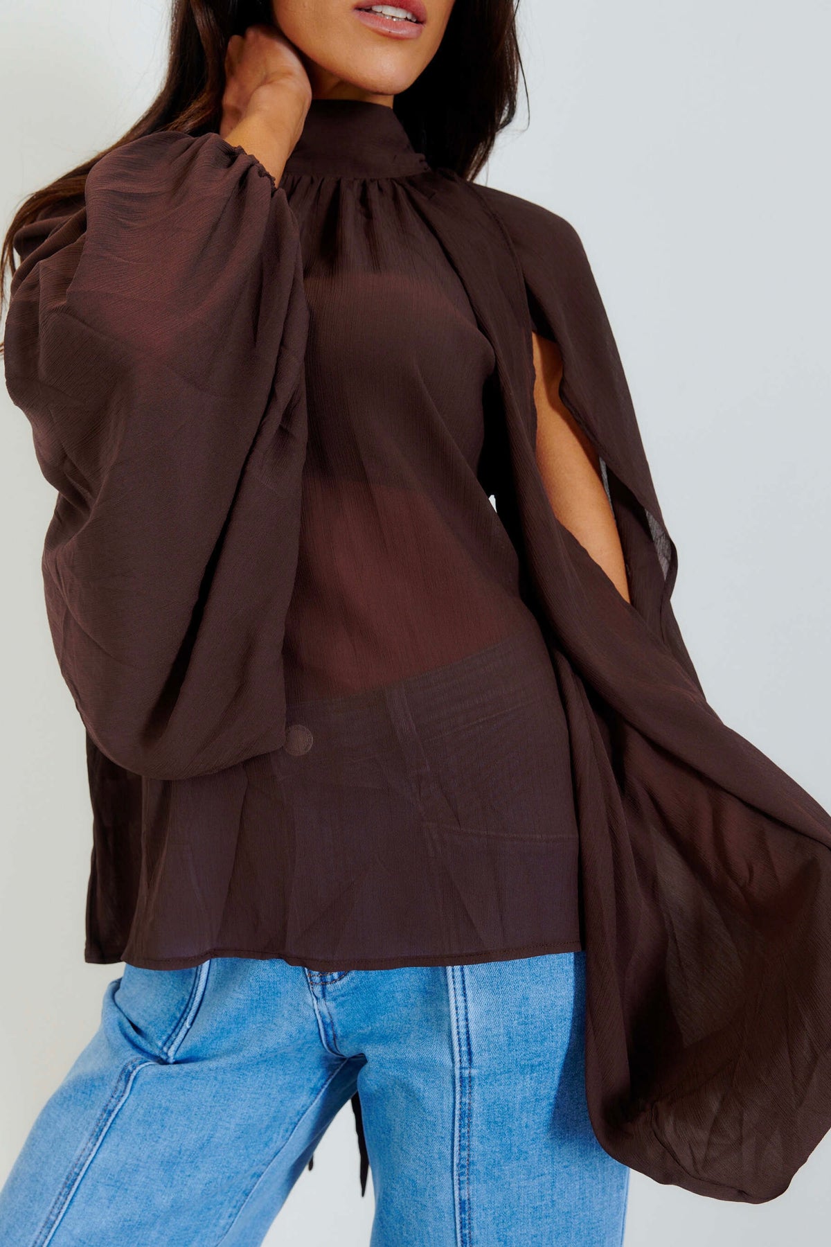 A woman wearing a dark brown sheer long-sleeve blouse with dramatic balloon sleeves and side cutouts, paired with light blue jeans, against a plain light gray background.