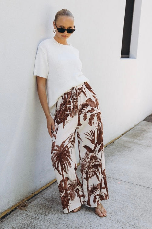 Person wearing a white t-shirt and brown palm leaf patterned pants against a white wall.