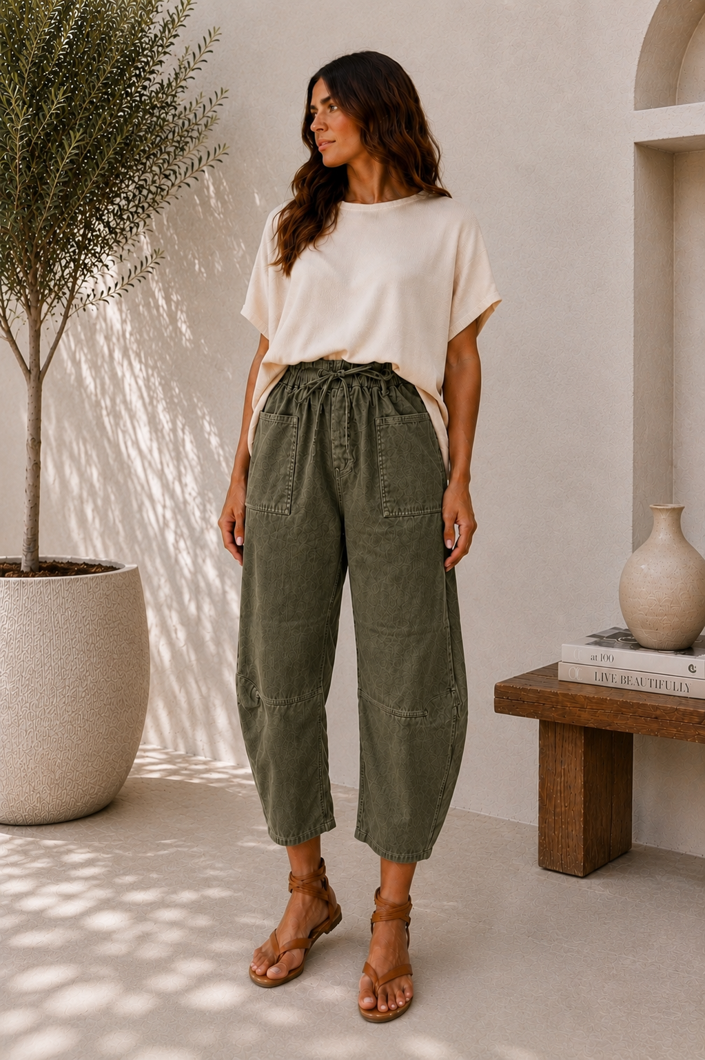 A woman stands in a bright, modern interior wearing a cream short-sleeve top and olive green drawstring cropped pants with brown strappy sandals.
