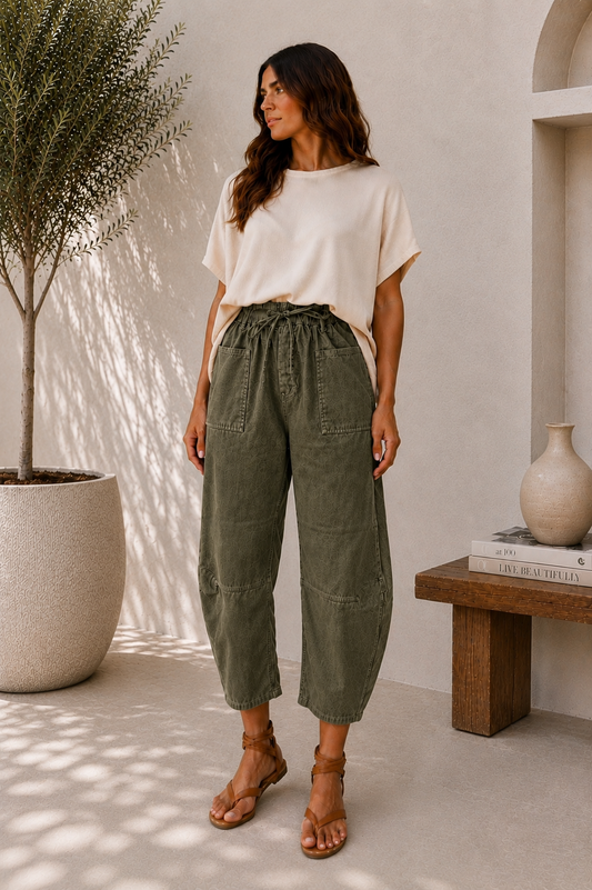 A woman stands in a bright, modern interior wearing a cream short-sleeve top and olive green drawstring cropped pants with brown strappy sandals.