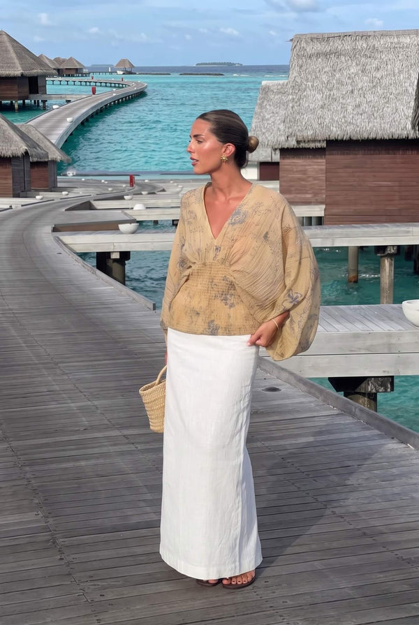 A woman stands on a wooden overwater walkway wearing a beige patterned blouse and a long white skirt, with turquoise water and thatched-roof bungalows in the background.
