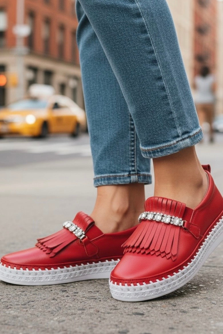Red leather slip-on shoes with fringe and rhinestone embellishments, worn with blue jeans on a city street.