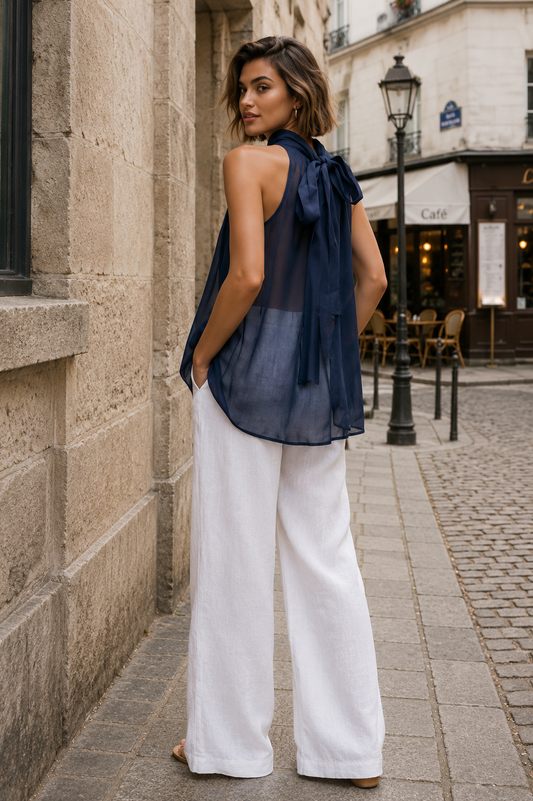 A woman stands on a cobblestone street wearing a sheer navy blue sleeveless top with a large bow detail at the neckline and white wide-leg pants, photographed in an outdoor urban setting.