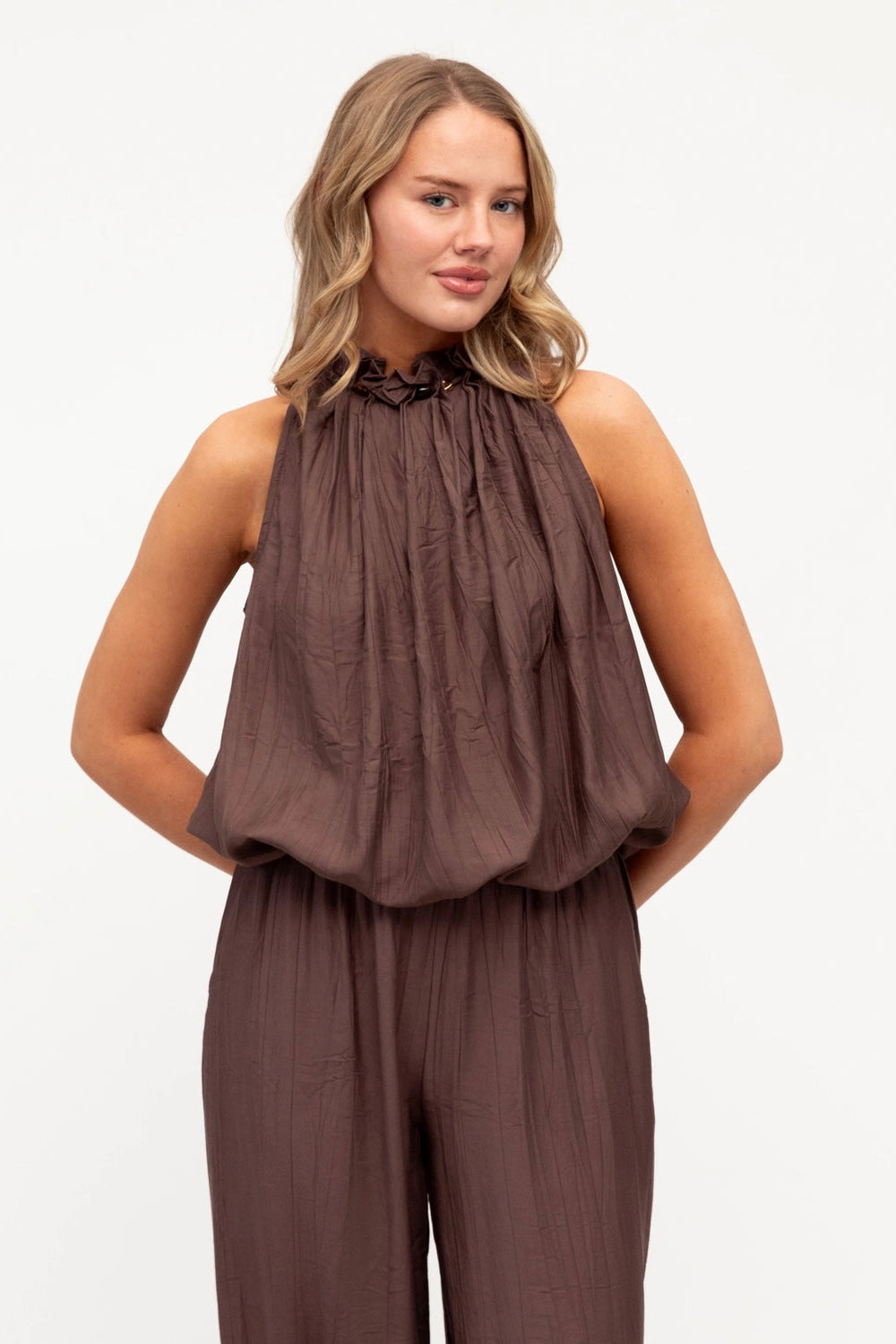 A woman modeling a sleeveless brown halter top and matching pleated pants against a plain white studio background.