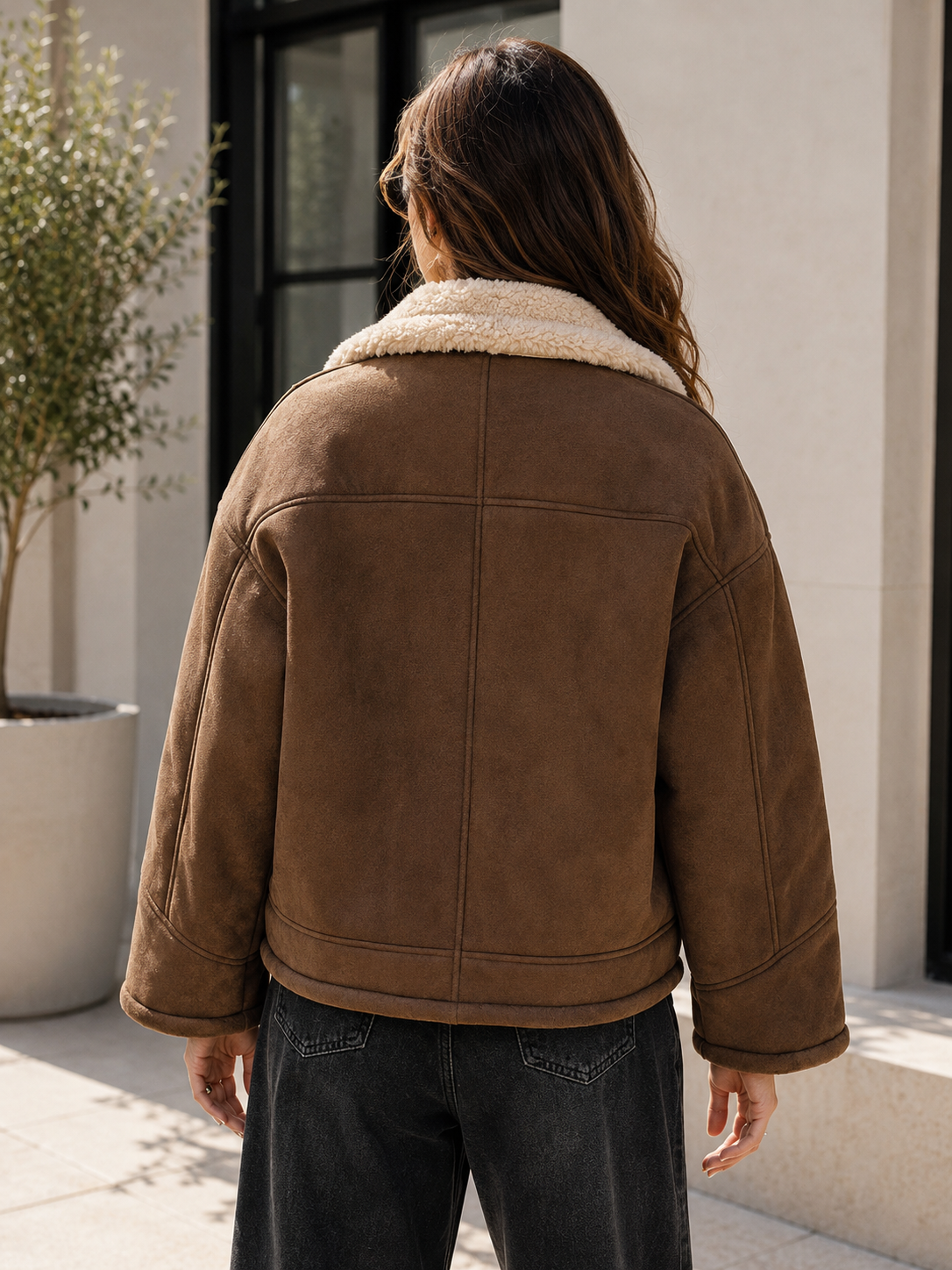 A person viewed from behind wearing a brown shearling-lined jacket with a thick cream-colored collar, paired with dark jeans, standing outdoors in front of a modern building.