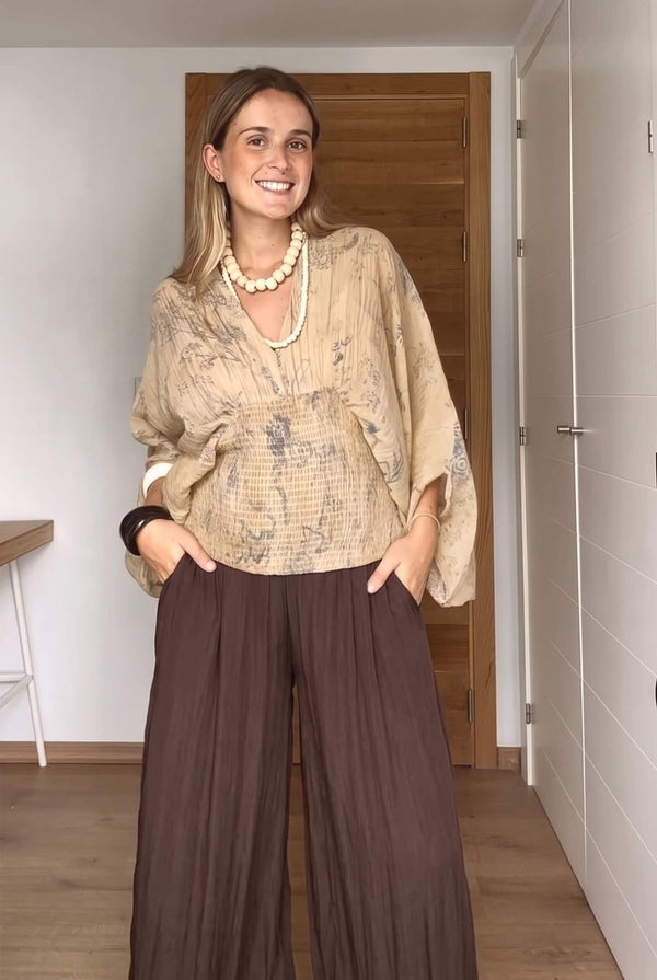 A woman modeling a beige patterned blouse with wide sleeves and dark brown wide-leg pleated pants, standing in an indoor room with a wooden door behind her.