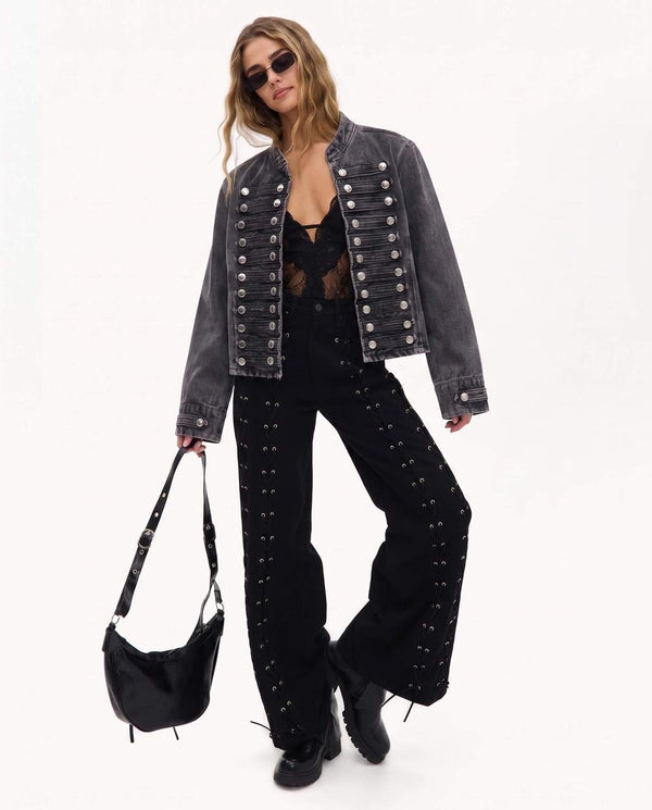 A model wearing a washed black denim jacket with silver studs, black studded wide-leg jeans, a black lace top, and holding a black crescent shoulder bag against a plain white background.