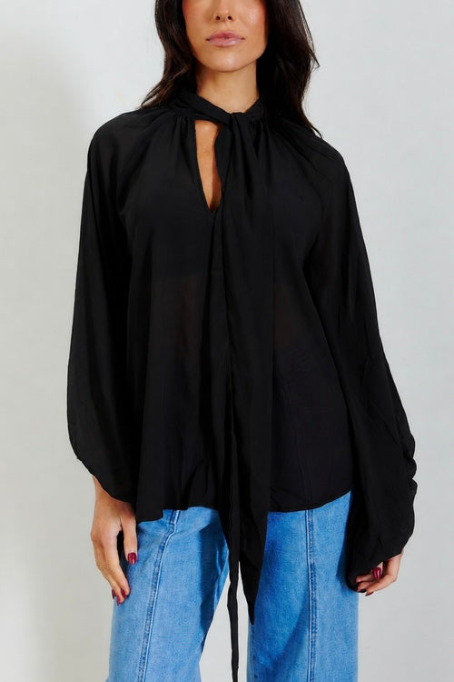A woman wearing a black long-sleeve blouse with a keyhole tie-neck detail and blue jeans, photographed against a plain light gray studio background.