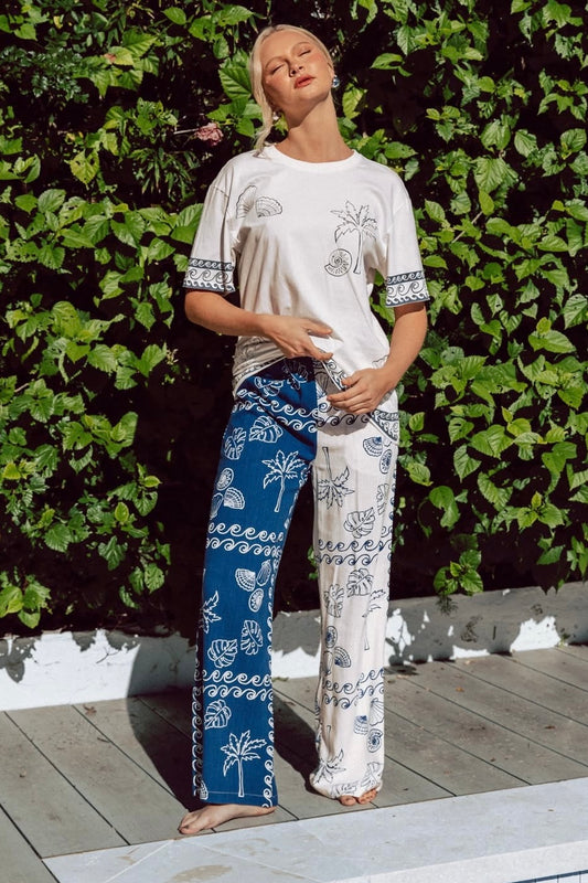 Person wearing a white t-shirt and blue and white patterned wide leg  pants standing against a green leafy background.