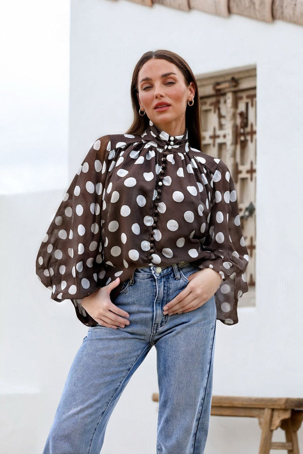 Woman wearing a brown polka dot blouse and blue jeans against a white wall.