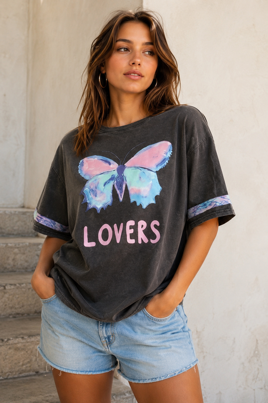 A woman wearing a dark gray washed graphic t-shirt with a colorful butterfly and the word 'LOVERS' in pink, paired with light blue denim shorts, standing against a light-colored wall.