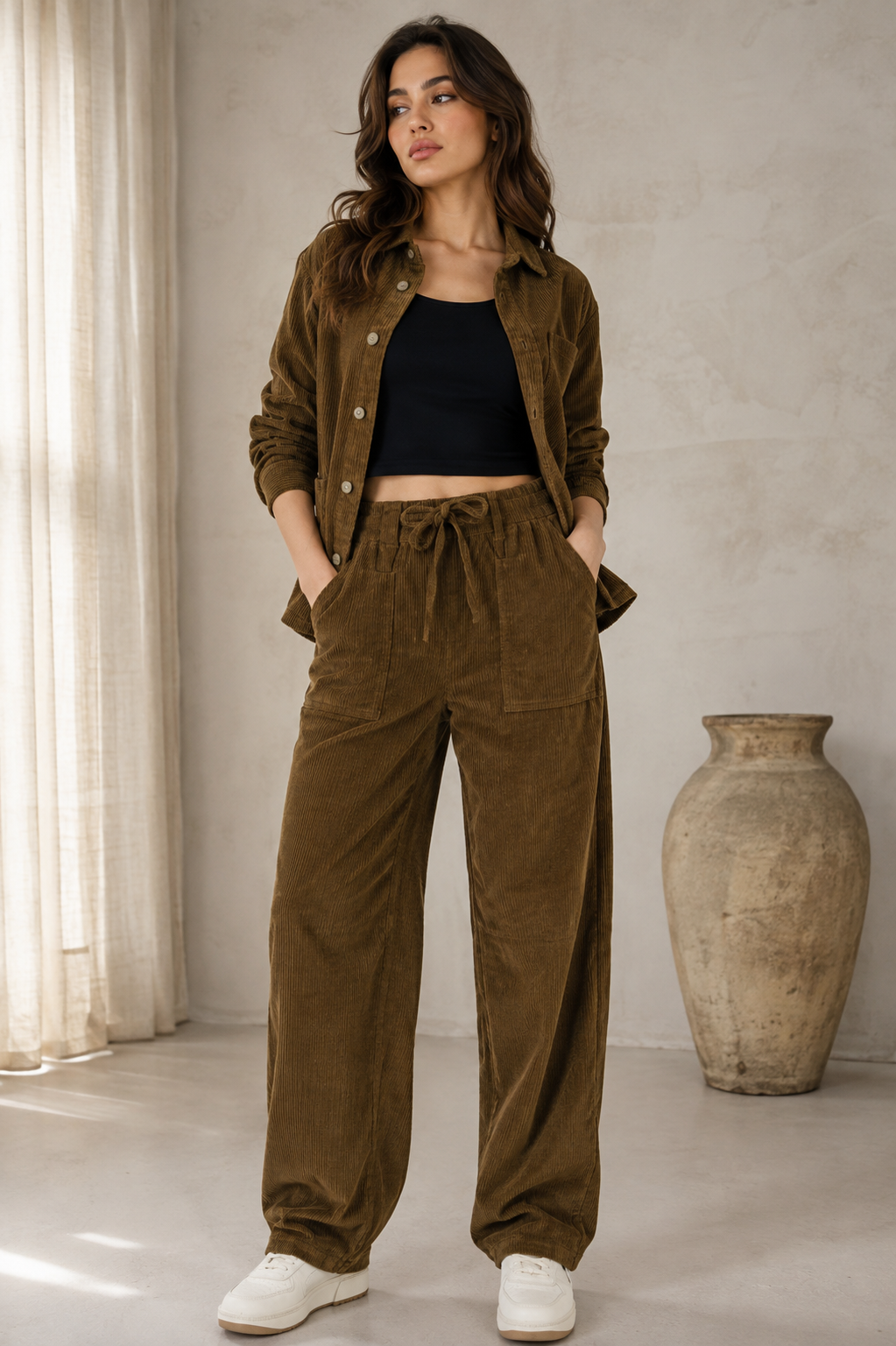 A woman modeling a pair of high-waisted, wide-leg brown corduroy pants with a drawstring waist, paired with a black crop top and white sneakers, against a plain light gray background.