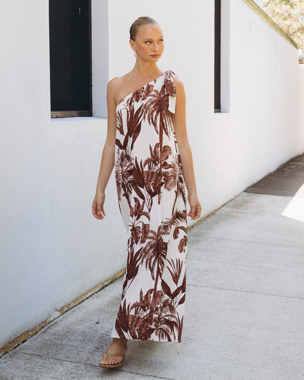 Woman wearing a one-shoulder floral dress standing on a sidewalk.