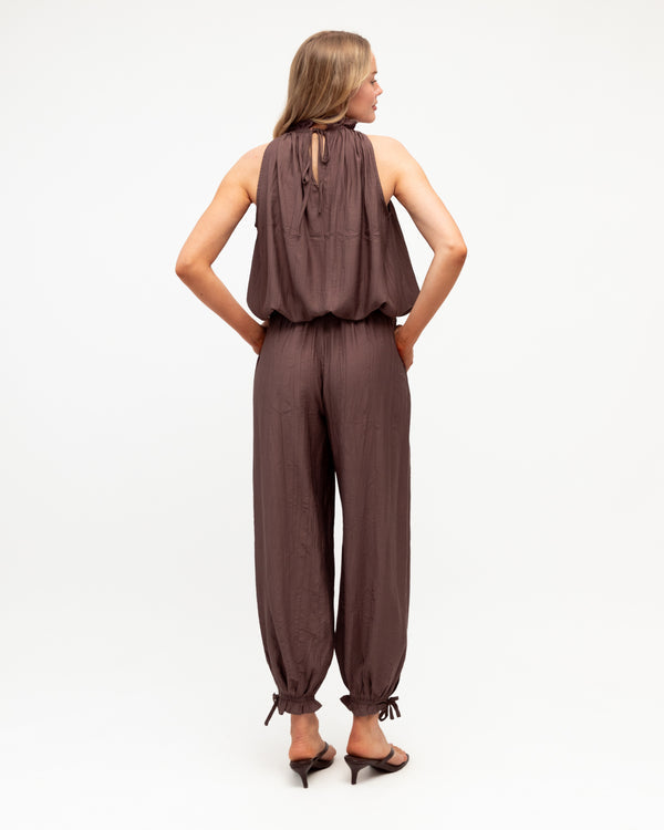 A woman viewed from the back wearing a sleeveless brown jumpsuit with a tie-back neckline and loose harem-style pants, standing against a plain white studio background.